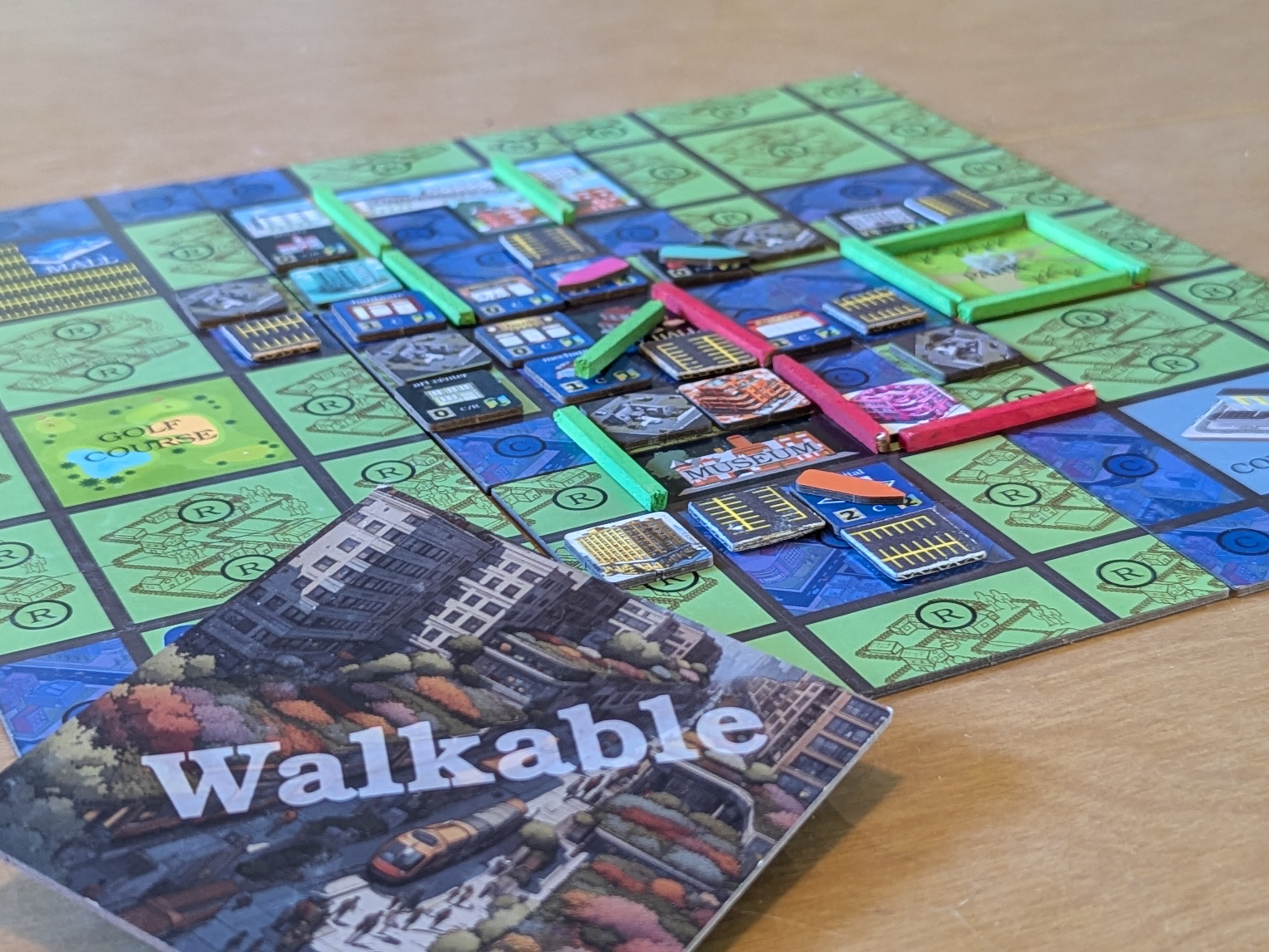 A game board with small city tiles on top of a gridded city board. In the bottom left there is an upside down board piece with the logo and a futuristic urban city with tram on it.